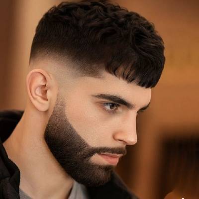 A man with a Caesar haircut featuring short, textured curls on top and a clean fade on the sides, complemented by a sharp beard and defined facial lines.