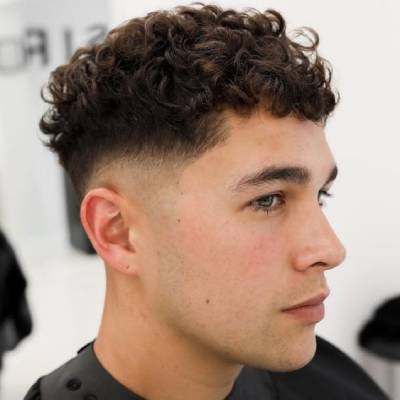 El hombre en la imagen tiene un corte de cabello estilo "crew cut" con rizos en la parte superior y un desvanecimiento en los lados.







