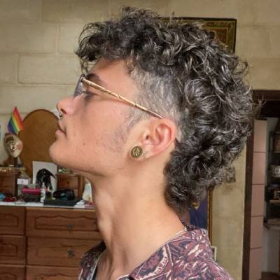 das Haar oben und hinten lockig bleibt und in einem natürlichen, voluminösen Stil getragen wird. Der Mann trägt eine Brille und Ohrringe und hat ein lässiges Outfit mit einem gemusterten Hemd.


