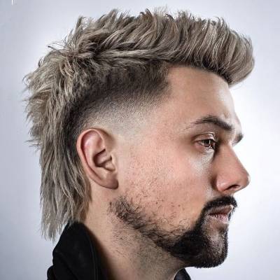 kurz geschnitten, während das Haar oben und am Hinterkopf länger ist und in einen spitzen Mohawk-Stil gestylt wurde. Der Mann hat einen gepflegten Bart und trägt ein schwarzes Oberteil.


