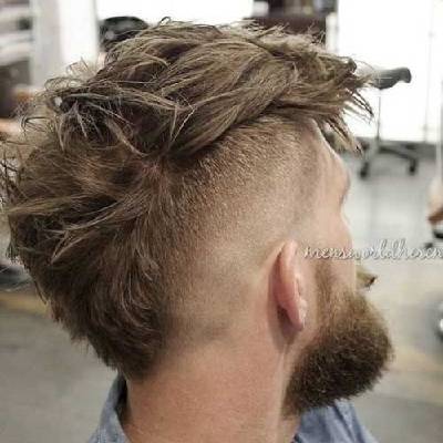 während das Haar oben voluminös und nach oben gestylt ist, was einen Faux-Hawk-Look erzeugt. Der Mann trägt einen Bart, und der Stil hat einen modernen, abenteuerlichen Charme.